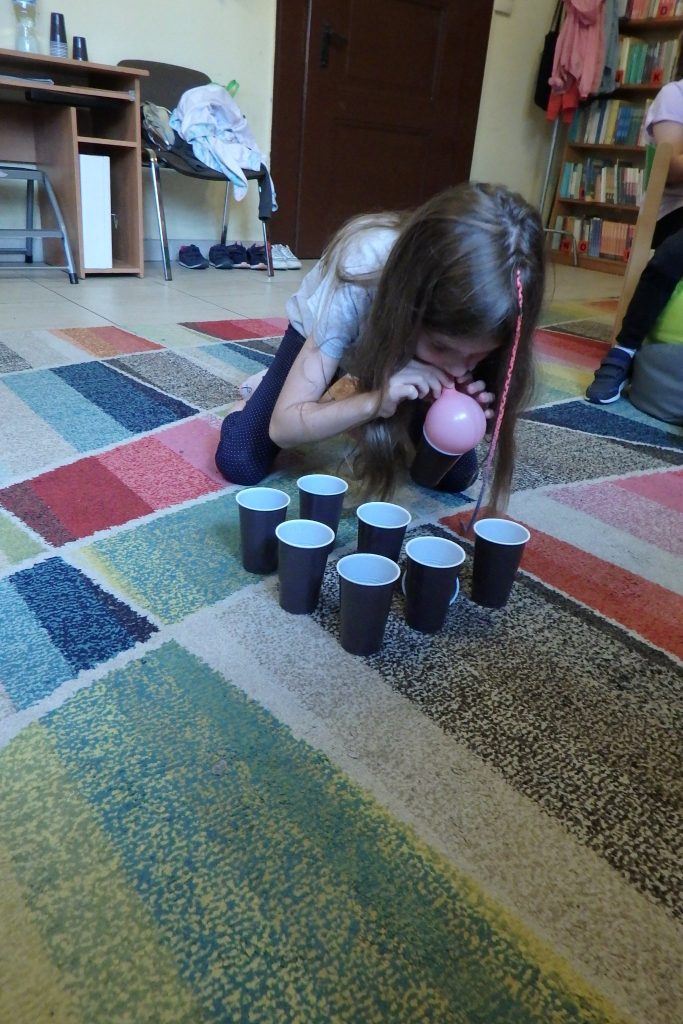 Na kolorowym dywanie kuca dziewczynka. Przed nią są ustawione w rzędach plastikowe kubeczki. Dziewczynka za pomocą balona składa kubki. W tle widać drzwi ,biurko i regał z książkami.