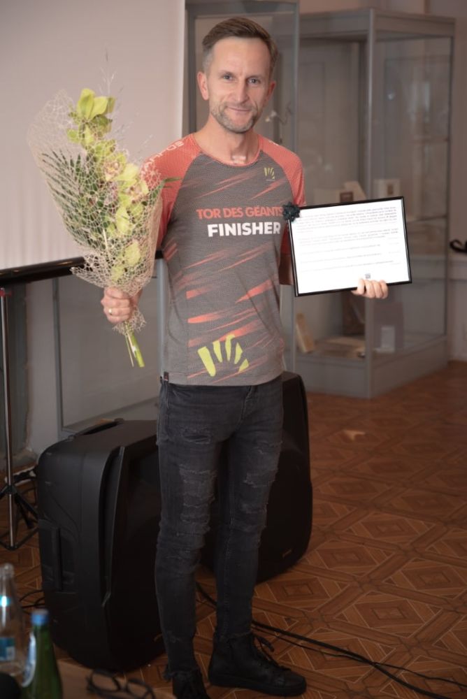 Mężczyzna pozujący do zdjęcia ubrany w sportową koszulkę z napisem TOR DES GEANTS FINISHER. W prawej ręce trzyma bukiet kwiatów. W lewej ręce ramkę ze zdjęciem.
