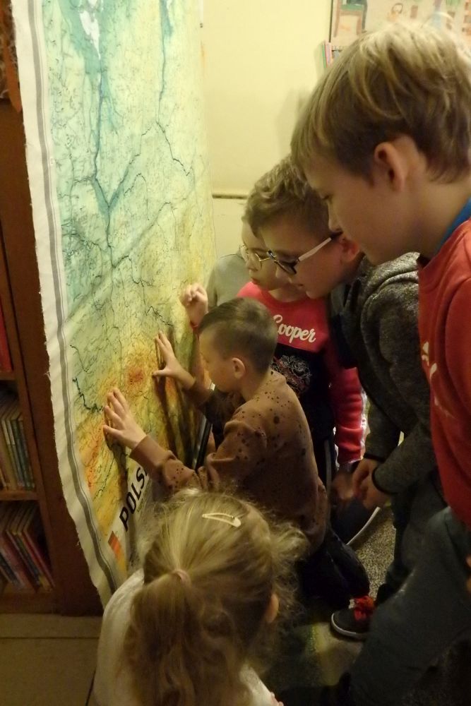 Z lewej strony duża mapa Polski. Z prawej strony grupa dzieci z zainteresowaniem obserwują mapę.