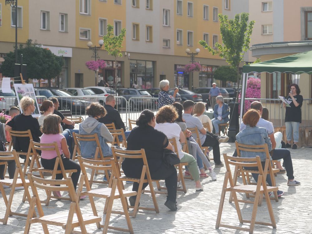 Grupa osób siedząca tyłem na krzesełkach w przestrzeni miejskiej.