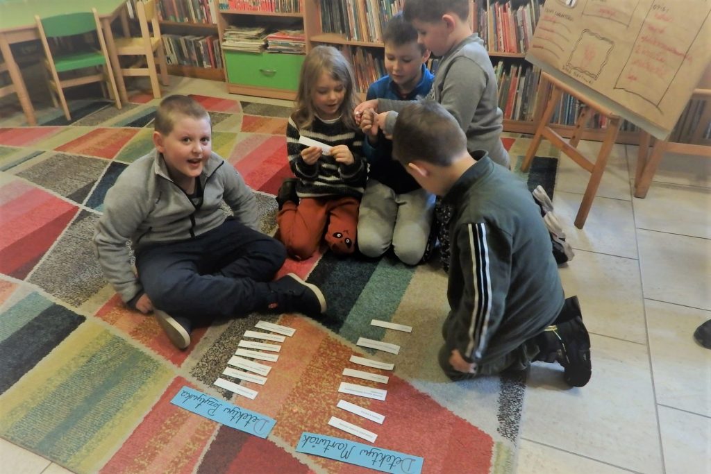 Na dywanie siedzi grupa dzieci. Przed nimi leżą kartoniki z zagadkami. W tle pomieszczenie biblioteki.