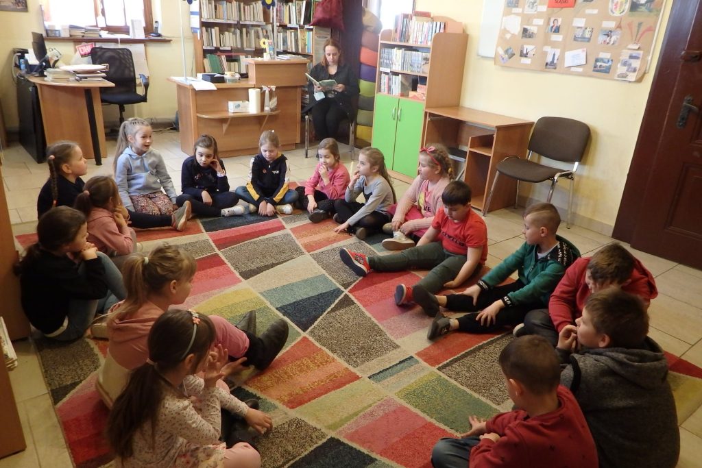 Wokół kolorowego dywanu siedzi grupa dzieci z klasy I. W głębi na krześle siedzi pani trzyma w rękach książkę, czyta. W tle pomieszczenie biblioteczne.