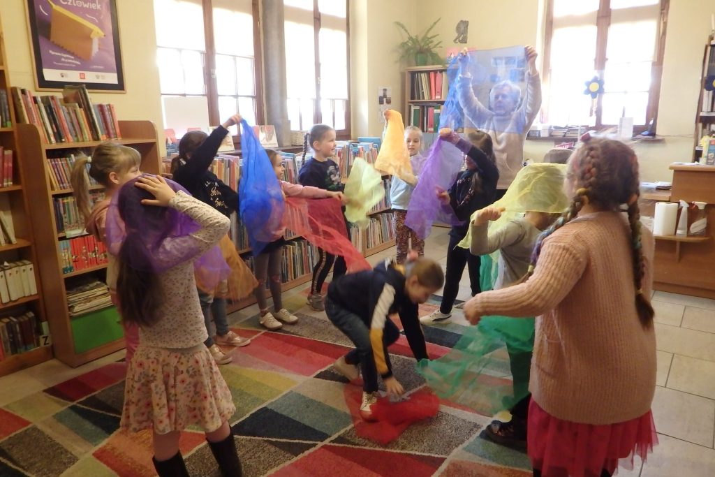 Na środku biblioteki grupa dzieci i dorosła pani trzymają nad głowami kolorowe, przezroczyste chusty, wykonują taniec kwiatów.