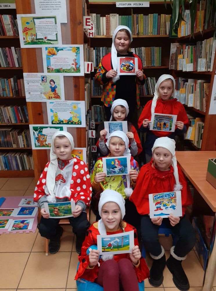 Grupa dzieci trzymających w dłoniach obrazki na tle regałów z książkami.