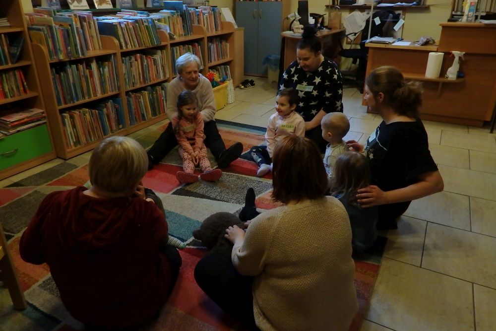 Na kolorowym dywanie siedzą w kole dzieci i ich opiekunowie, w centrum prowadząca zajęcia bibliotekarka, prezentuje masażyki relaksacyjne. Panie wykonują masażyki na plecach swoich dzieci. W tle od lewej strony regał z książkami i lady biblioteczne.