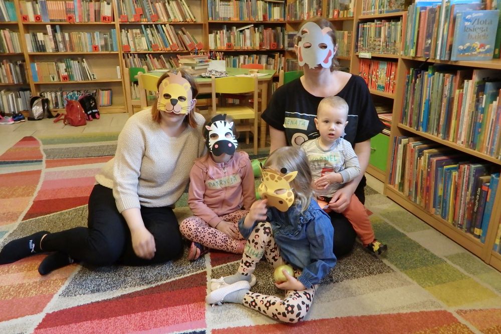 Na kolorowym dywanie siedzi 3 dzieci i 2 panie. Wszyscy (oprócz jednego małego dziecka) mają na twarzach maski zwierząt. W tle regały z książkami.