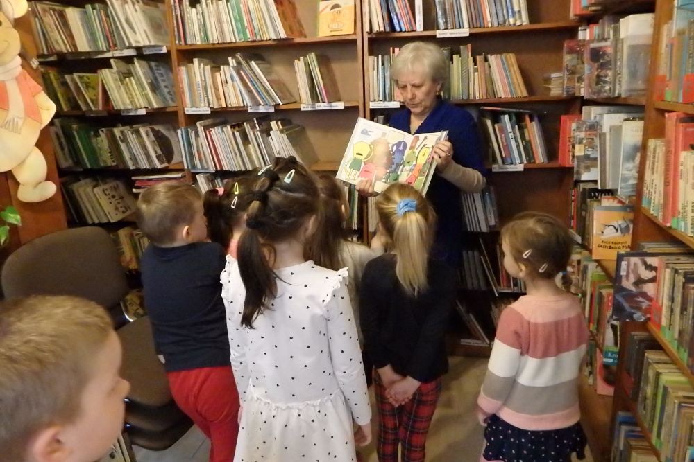 Na pierwszym planie widać grupę dzieci stojących tyłem. Patrzą na siwą bibliotekarkę w niebieskim swetrze, trzymającą w rękach otwartą, kolorową książkę. W tle regały z książkami.