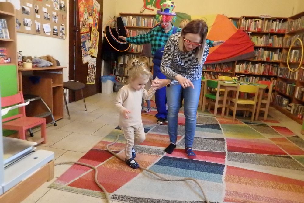 Na kolorowym dywanie rozciągnięta gruba lina po której idzie dziewczynka w wieku 2 lat, obok idzie mama i trzyma dziewczynkę za rękę. Za nimi po linie idzie kolorowy klaun i inne słabo widoczne osoby. W tle regały z książkami, po lewej stronie drzwi.