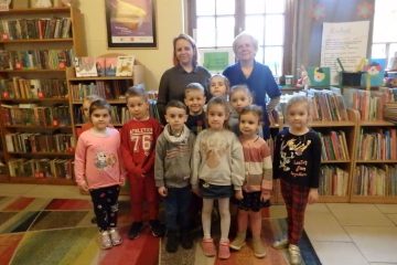 W centrum zdjęcia grupa przedszkolaków stoi w dwóch rzędach. Za nimi 2 bibliotekarki. W tle okno i niskie regały z książkami.