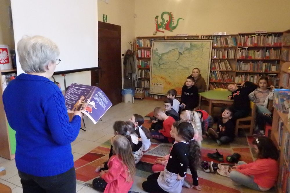 Z lewej strony stoi tyłem siwa bibliotekarka w niebieskim swetrze, trzyma w rękach czasopismo. Na środku kolorowego dywanu siedzi grupa dzieci, z tyłu przy zielonym stoliku siedzi kilka osób, w tle wisi mapa Polski.