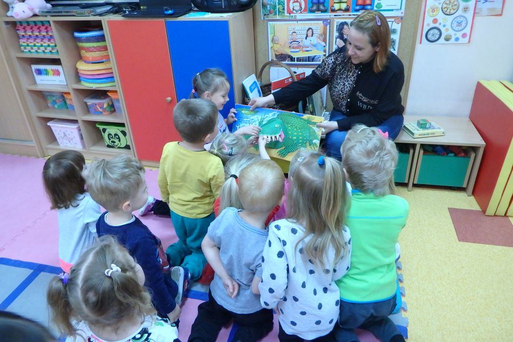 Grupa dzieci siedzi tyłem na kolorowej podłodze. Bibliotekarka pokazuje im książkę z dużym, rozkładanym krokodylem. W tle stoją meble z kolorowymi zabawkami.