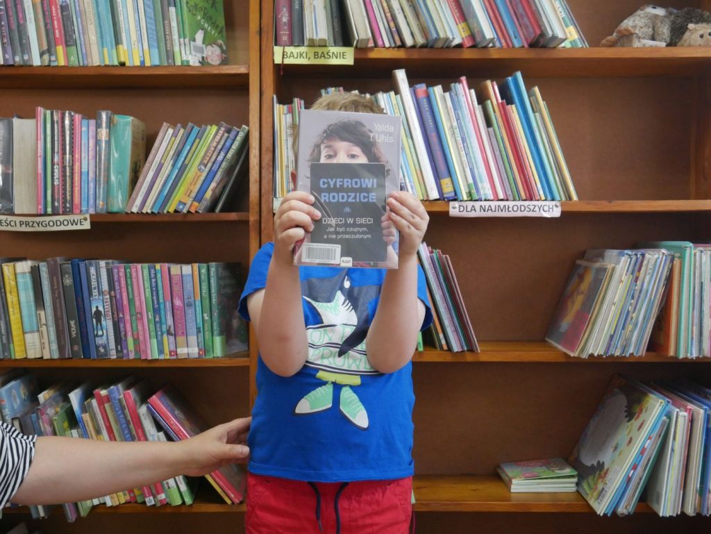 Stojący chłopiec zasłaniający twarz książką na tle regałów z książkami