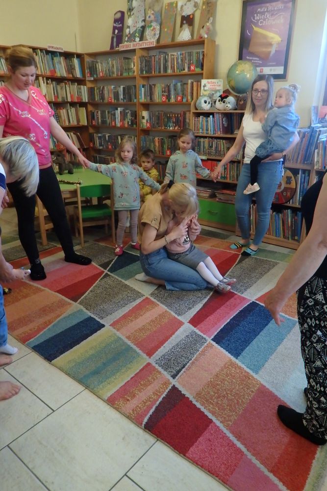 Na kolorowym dywanie siedzi mama z dzieckiem, które zasłaniają oczy dłońmi. Wokół są dorośli z dziećmi trzymające się za ręce. W tle znajdują się regały z książkami.