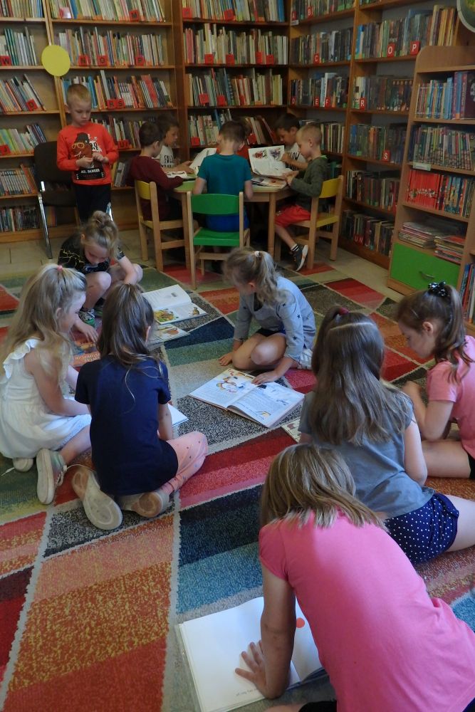 Na kolorowym dywanie siedzą dziewczynki i oglądają książki. W dalszej części, przy zielonym stole siedzą chłopcy, którzy również oglądają książki. W tle są regały z książkami.