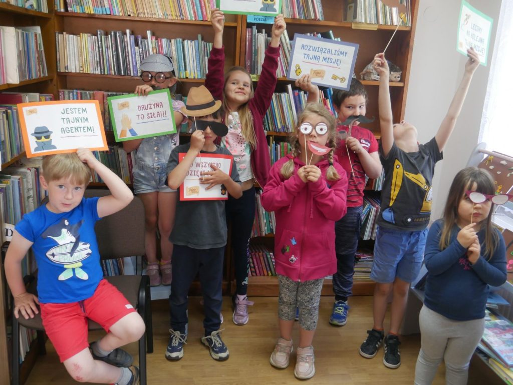 Grupa dzieci trzymających maski i kartki na tle regałów z książkami