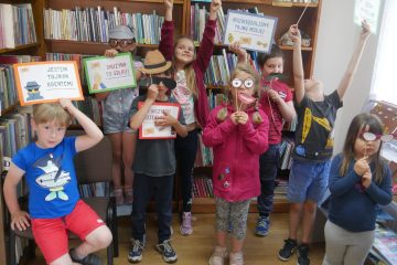 Grupa dzieci trzymających maski i kartki na tle regałów z książkami