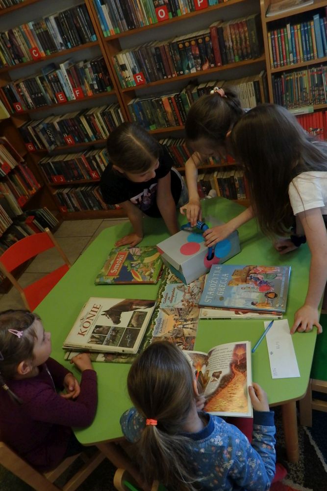 Na zielonym stole widać ręce dzieci, które z rozrzuconych elementów układają obrazek. Z boku leżą książki.