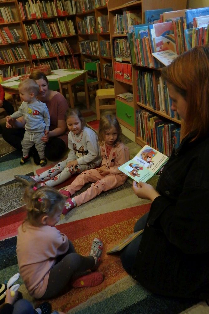 Po prawej stronie siedzi bibliotekarka i trzyma książkę. Na kolorowym dywanie siedzą dzieci i słuchają. W tle stoi stolik i regały z książkami.
