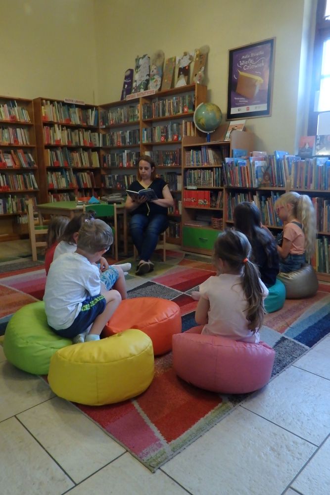 Na pierwszym planie tyłem siedzą dzieci na kolorowych pufach. Przed nimi siedzi pani bibliotekarka ,w rękach trzyma książkę. Obok stoi zielony stolik. W tle regały z książkami.