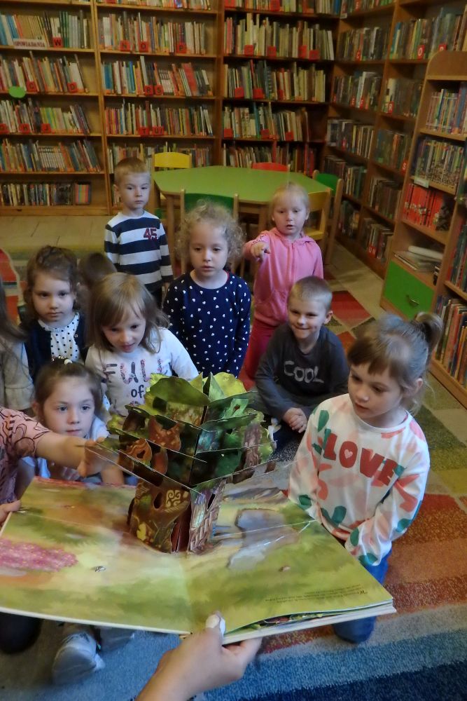 Na kolorowym dywanie siedzi grupa dzieci. Dzieci oglądają książkę w formie rozkładanego drzewa, która jest przed nimi.