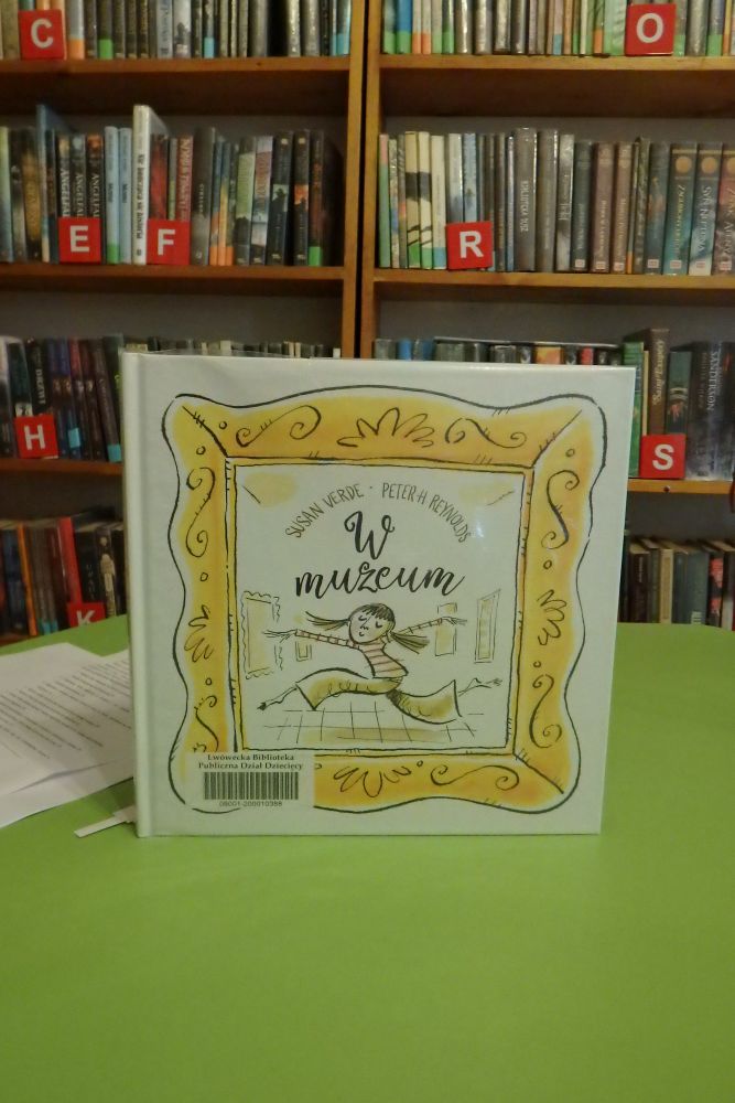 Na środku zielonego stolika jest postawiona książka pt. „W muzeum” autorstwa Susan Verde. W tle regały z książkami.
