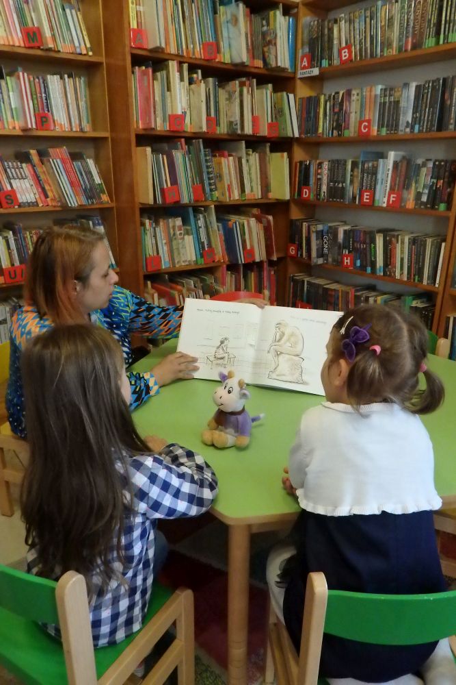 Na krzesełkach wokół zielonego stolika siedzą dwie dziewczynki i pani bibliotekarka. Bibliotekarka pokazuje dziewczynkom książkę z ilustracjami. Na środku stołu leży mały miś. W tle regały z książkami.