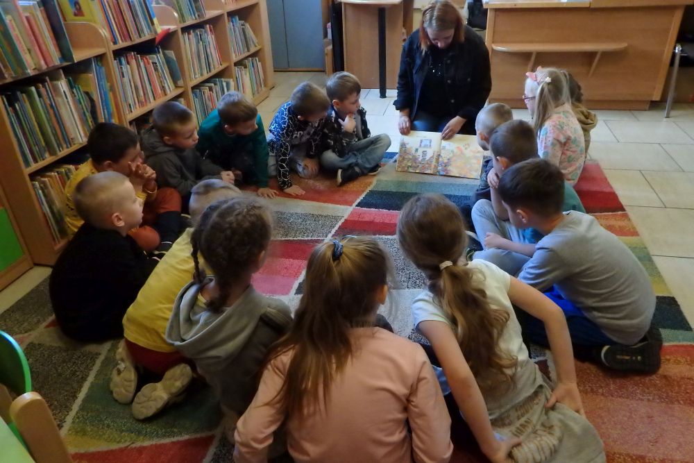 Na kolorowym dywanie siedzi grupa dzieci a bibliotekarka czyta oraz pokazuje im książkę. Za bibliotekarką są biurka a po lewej stronie regały z książkami.
