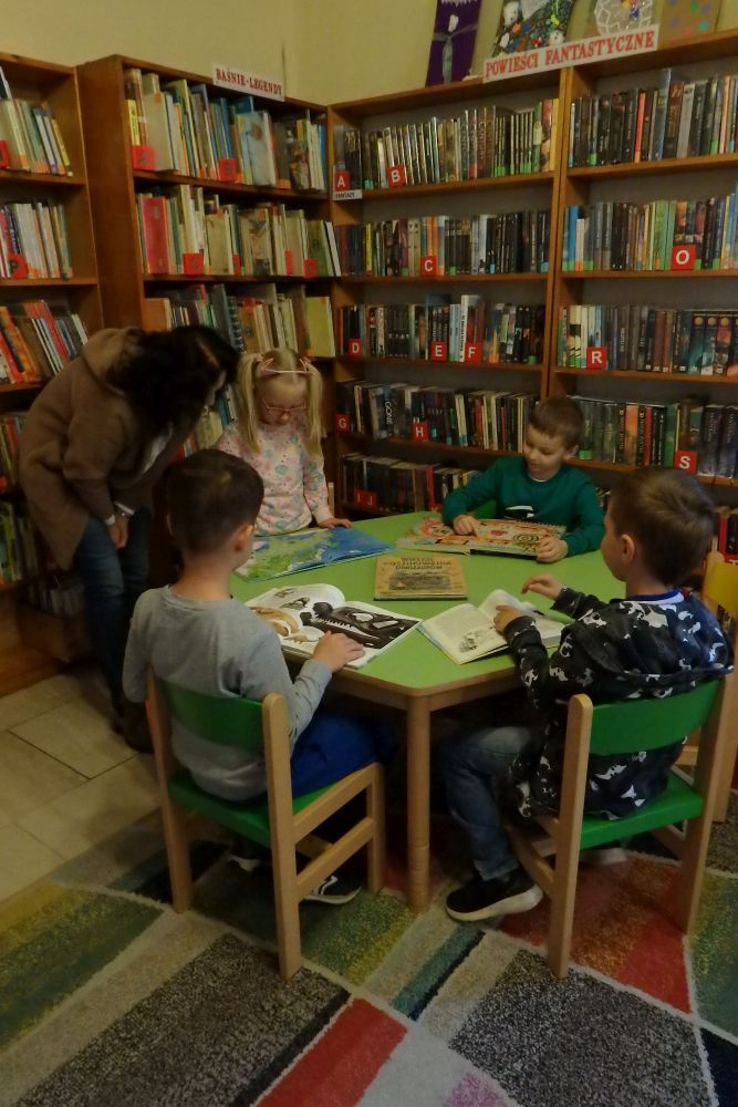 Przy zielonym stole siedzą dzieci i oglądają książki. Jedna dziewczynka stoi i pokazuje coś pani nauczycielce. W tle stoją regały z książkami.