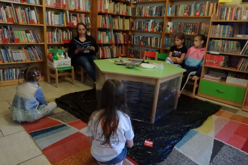 Na środku stoi stół przy którym siedzi pani bibliotekarka i dwie dziewczynki. Na stole leżą pędzle i szare gazety, a pod nim rozłożona jest czarna folia. Na dywanie siedzą tyłem dwie dziewczynki i słuchają .Pani bibliotekarka trzyma w ręku książkę ,czyta. Obok soi krzesło na którym jest pudełko z farbami.