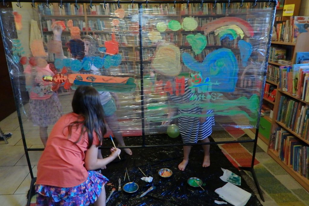 Na fotografii znajduje się ogromny stojak owinięty folia spożywczą. Na kolorowym dywanie leży czarna folia, na której stoją dzieci i malują kolorowymi farbami po rozciągniętej folii spożywczej. Wokół stoją regały z książkami.