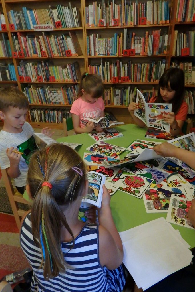 Na zielonym stoliku leżą komiksy „Tytus, Romek i A’Tomek”. Wokół stolika siedzą dzieci na zielonych krzesełkach i przeglądają komiksy. W tle stoją regały z książkami.