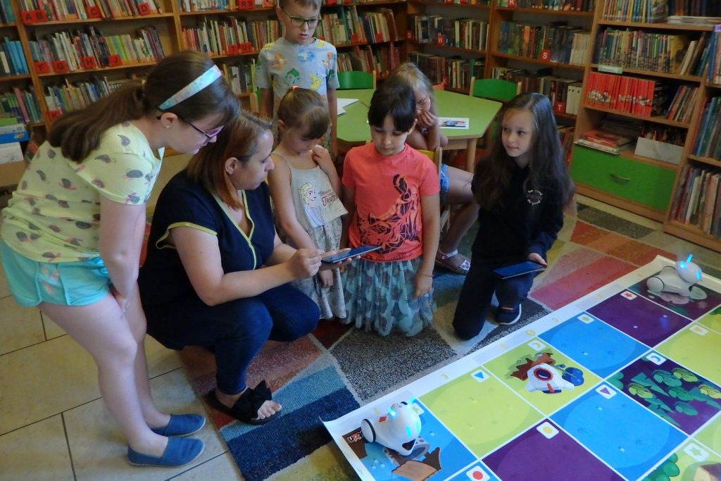 Grupa dzieci wraz z panią bibliotekarką siedzą na kolorowym dywanie. Pani bibliotekarka i jedno z dzieci trzymają tablety. Przed nimi leży kolorowa mata, na której leżą photony. W tle stoi zielony stolik oraz regały z książkami.