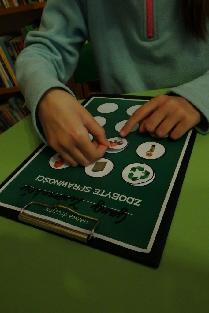 Na zielonym stoliku leży karta na sprawności z przyklejonymi symbolami zdobytych sprawności. Dziecko przykleja kolejną naklejką.