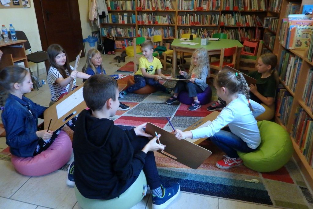 Na kolorowym dywanie w kole siedzi grupa dzieci. Podają sobie podkładki z kartkami. W tle są regały z książkami.