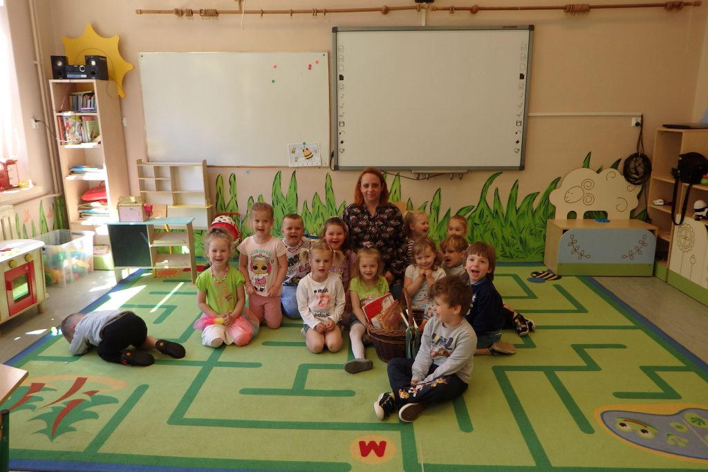Na zielonym dywanie siedzi grupa przedszkolaków wraz z Panią bibliotekarką. Za nimi widać ścianę, na której jest ręcznie namalowana zielona trwa, wiszą również dwie tablice. Po bokach widać regały z zabawkami