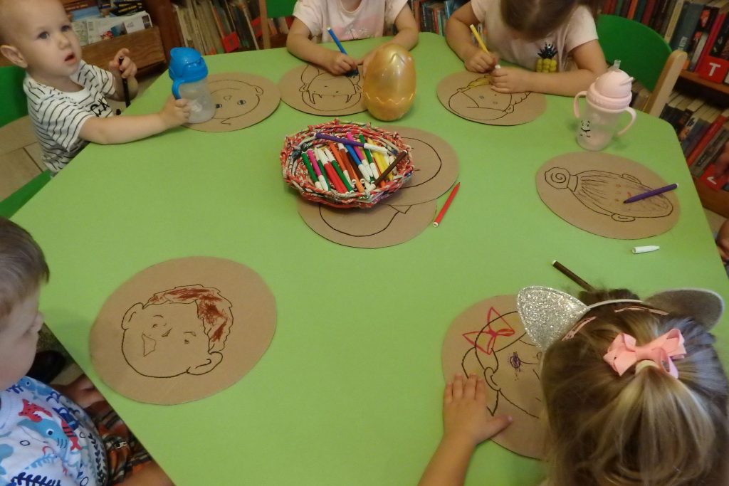 Przy zielonym stoliku siedzą dzieci, które na tekturowym kołach rysują swoje autoportrety. Na środku stolika leż papiery koszyk z kolorowymi kredkami.