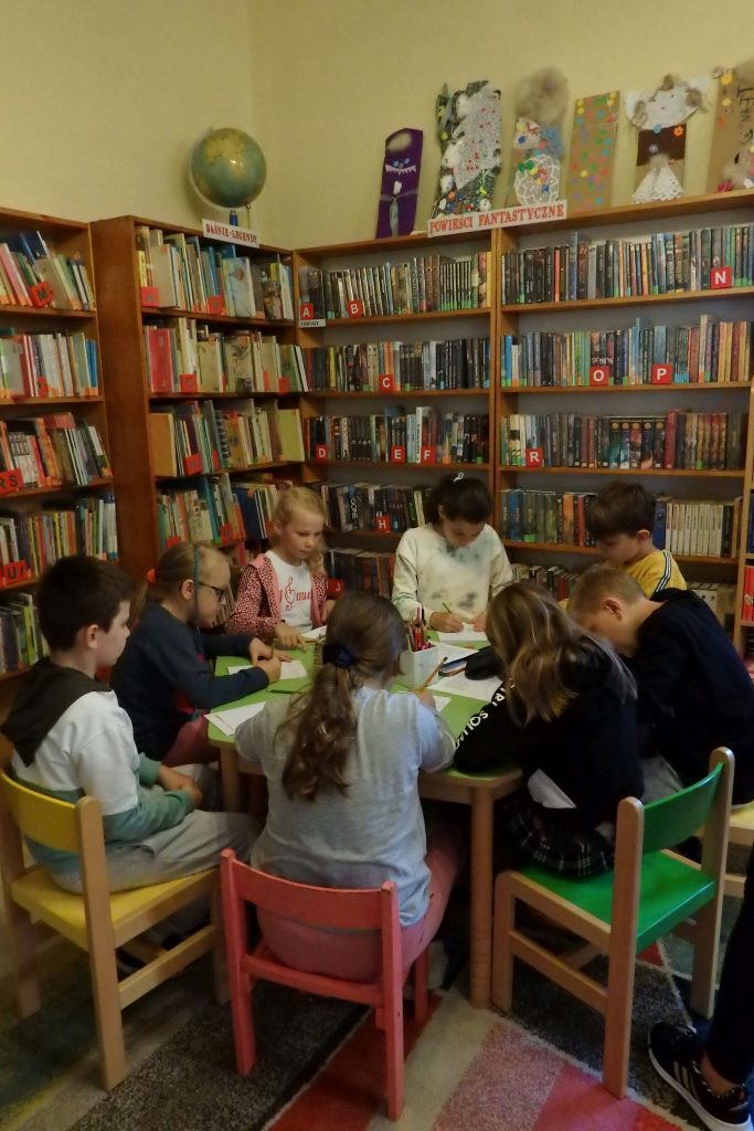 Przy zielonym stoliku na kolorowych krzesełkach siedzi grupa dzieci, które rysują. W tle stoja regały z kolorowymi książkami.