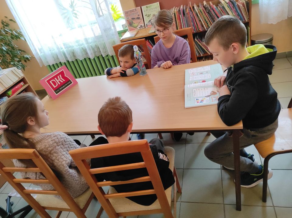 Przy stole siedzą dwie dziewczynki i trzech chłopców. Jeden z nich czyta książkę. Na stole stoi różowe logo Dyskusyjnych Klubów Książki. W tle jest regał z książkami i okno.