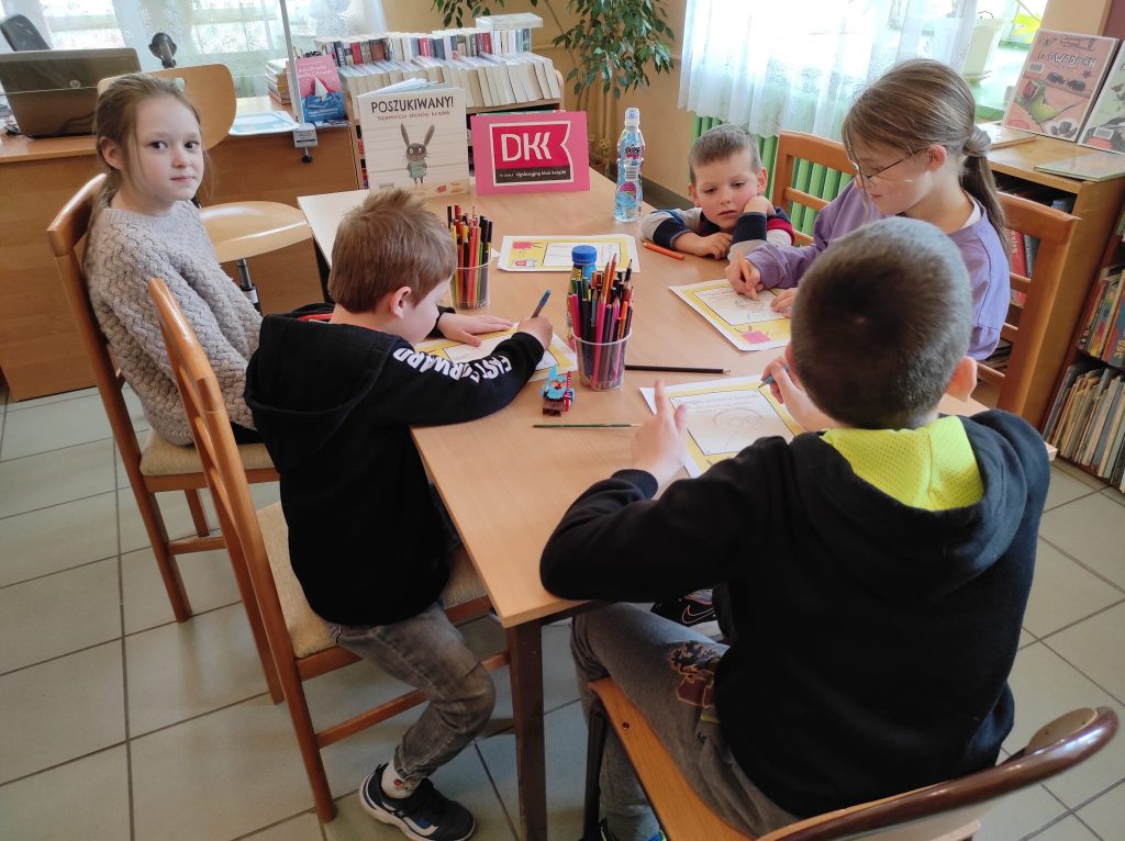 2. Przy stole siedzi pięcioro dzieci, dwie dziewczynki i trzech chłopców. Na stole są kolorowe kartki oraz kredki. Dzieci rysują ludzkie postacie.