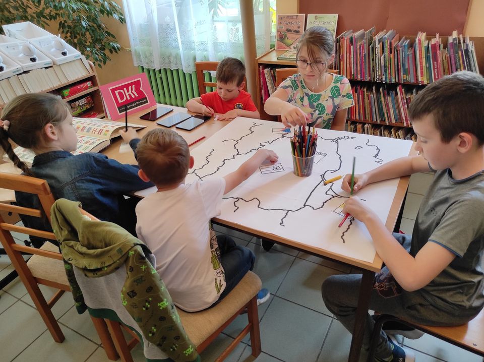 Przy stole siedzi trzech chłopców i dwie dziewczynki. Na stole leży kolorowanka z mapą Polski i sąsiednimi państwami oraz kredki.