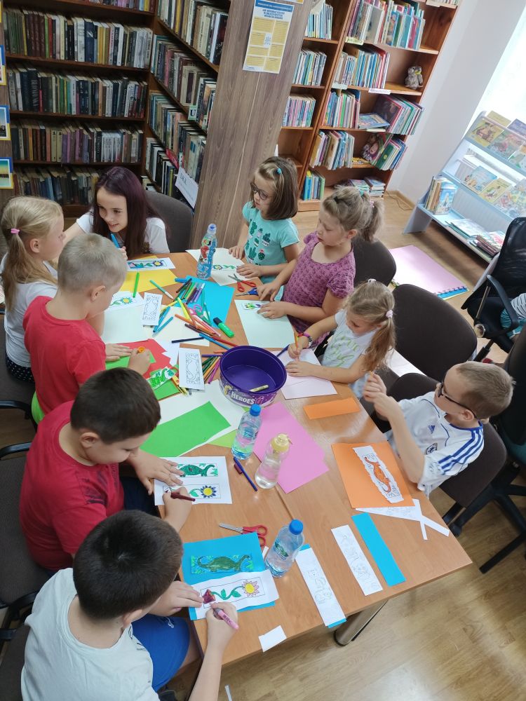 Grupa dzieci siedzących przy stole, wykonująca prace plastyczne. W tle regały z książkami.