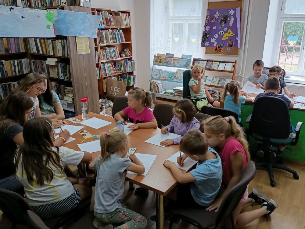 . Dwie grupy dzieci siedzących przy stołach i rysujących obrazki. W tle, z lewej strony, regały z książkami. Z prawej okna.