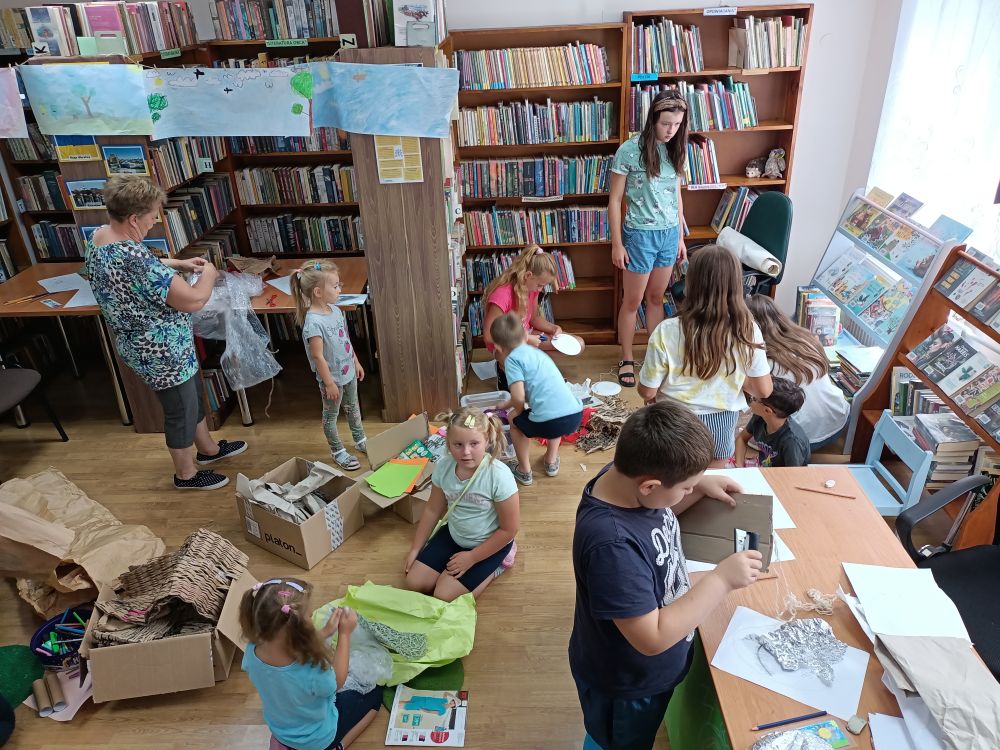 Grupa dzieci w pomieszczeniu biblioteki, wykonujących papierowe stroje. W tle regały z książkami.
