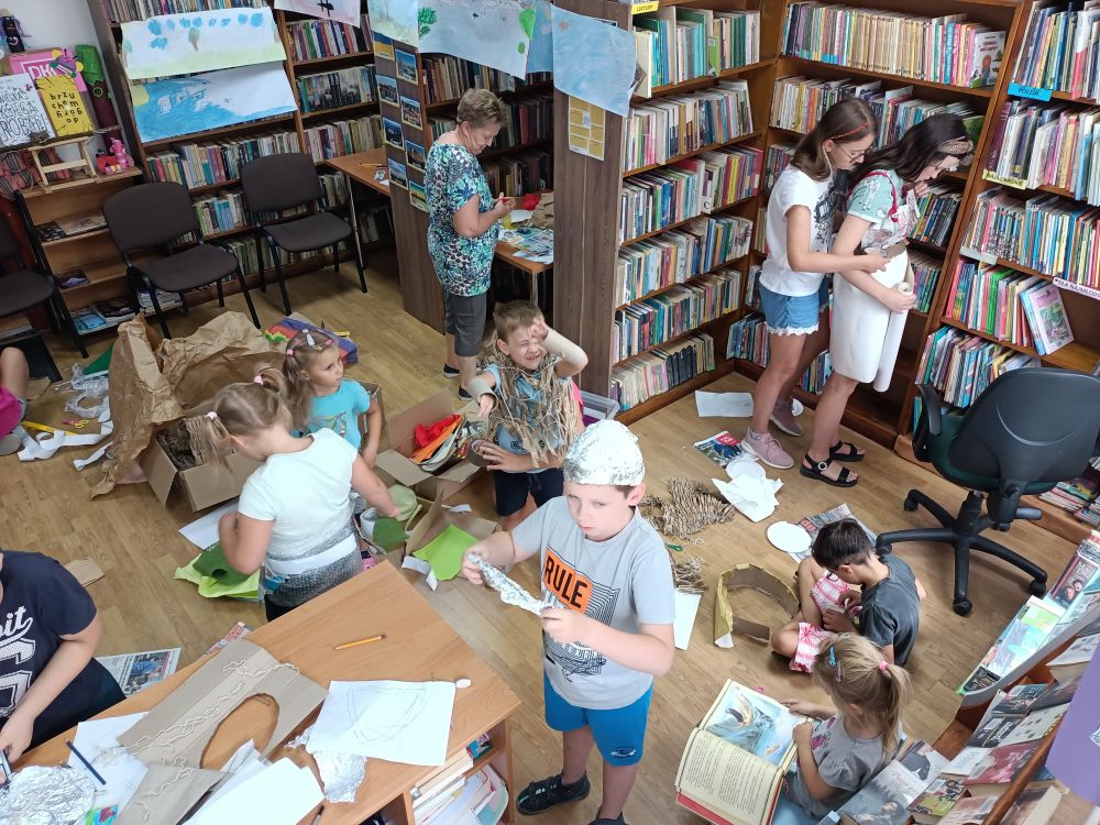 Grupa dzieci w pomieszczeniu biblioteki, wykonujących papierowe stroje. W tle regały z książkami.