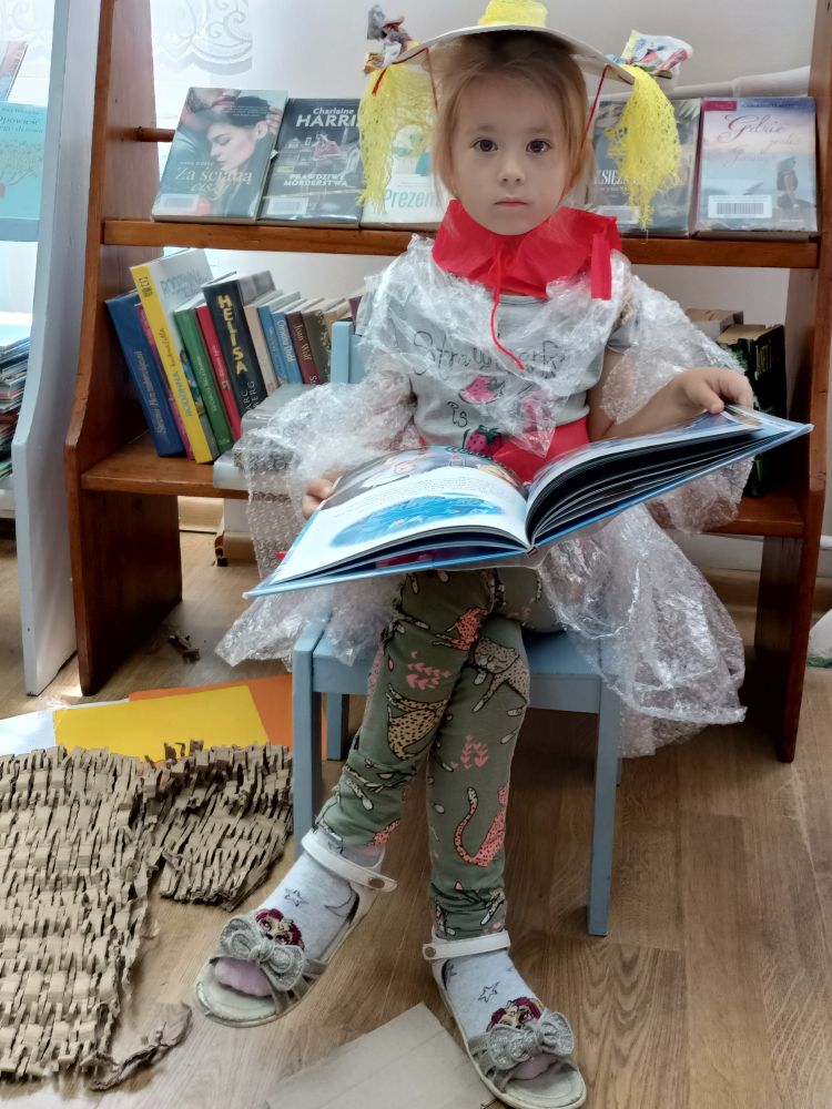 Mała dziewczynka siedząca na krzesełku, ubrana w kapelusz i sukienkę z folii i oglądająca książkę. Za nią regał z książkami.