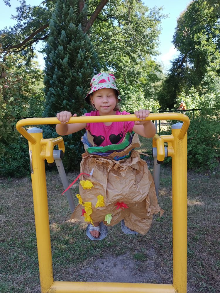 Dziewczynka ubrana w papierową sukienkę i huśtająca się na urządzeniu do ćwiczeń. W tle drzewa.