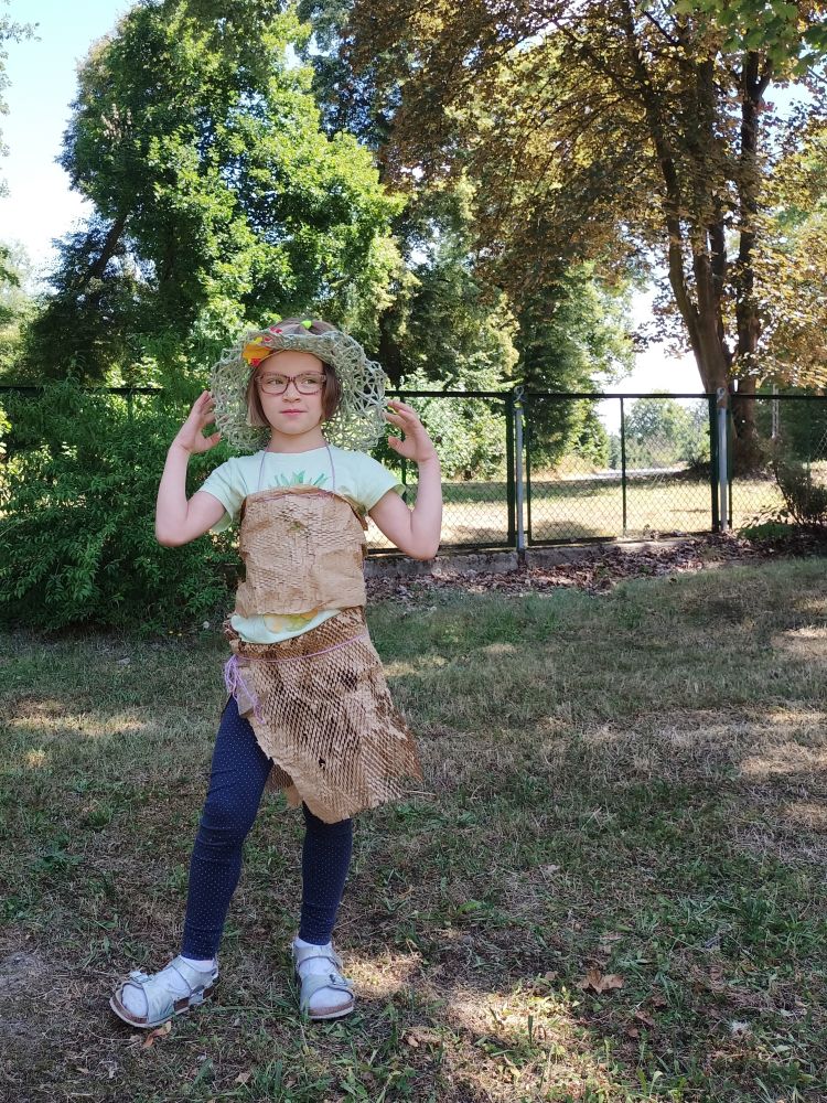 Dziewczynka stojąca na dworze, ubrana w papierową bluzeczkę i spódniczkę. Na głowie kapelusz, który podtrzymuje dłońmi. W tle trawa, ogrodzenie z siatki i drzewa.