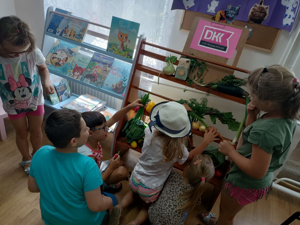 Grupa dzieci przy małym regale z warzywami i logiem Dyskusyjnego Klubu Książki.
