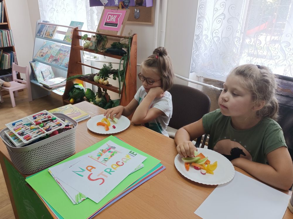 Dwie dziewczynki siedzące przy stole i jedzące warzywa z talerzyków. W tle mały regał z warzywami i logiem Dyskusyjnego Klubu Książki oraz mały regał z książkami.