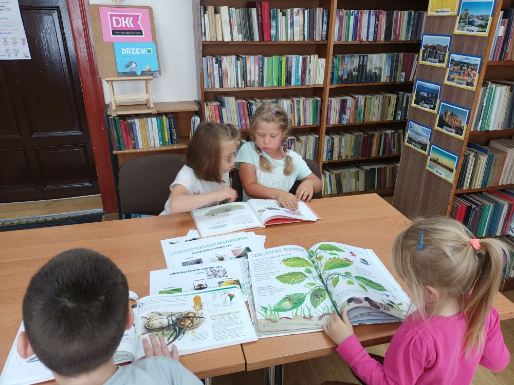 Czworo dzieci siedzi przy stole i ogląda książki o owadach. W tle, z prawej strony, drzwi. Z lewej regały z książkami.